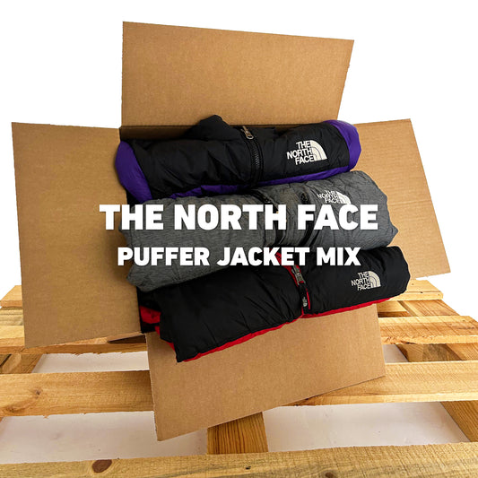The North Face Puffer Jacket Mix | Vintagebulk met drie opgevouwen jassen in een open kartonnen doos op een houten pallet, ideaal voor groothandel.
